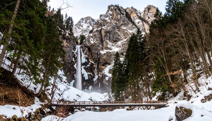 A magnificent winter waterfall cascading down a snowy mountainside, framed by towering evergreen trees and a wooden footbridge.