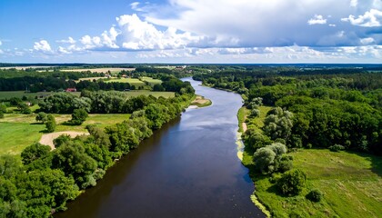 Obraz premium Serene river winds through a landscape of lush greenery and farmlands under a partly cloudy sky.