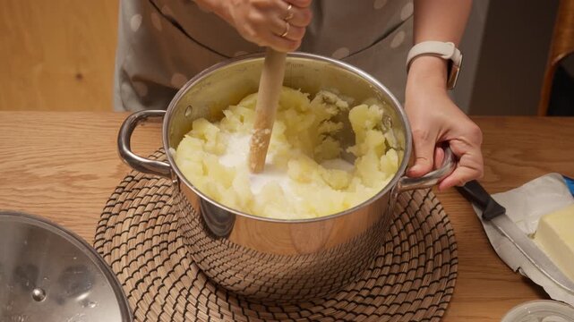 A cook adds milk from a glass cup to the pot of mashed potatoes to achieve a smooth and creamy texture. She continues to mix with a masher, preparing a delicious homemade side dish.