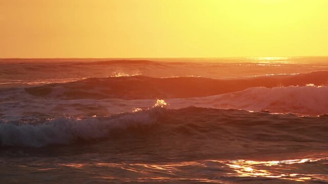 Ocean surf at dawn, waves close-up. Background, man surfing. Sky is golden, yellow-orange and beautifully reflected in water