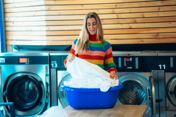 Smiling woman stands with blue basket in laundromat after washing clothes surrounded by modern self...