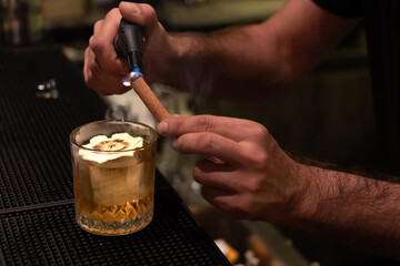 A bartender uses a culinary torch preparing a golden cocktail served in textured glass with large ice cube