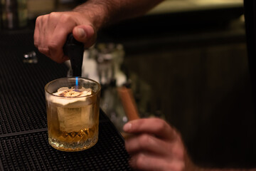 A bartender uses a culinary torch preparing a golden cocktail served in textured glass with large ice cube