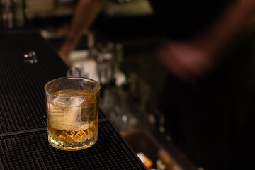 Freshly poured, golden cocktail with a large, square ice cube in a glass on black rubber bar mat with blurred bartender in background