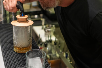 A bartender uses a culinary torch to add smoke to cocktail in a tall, textured glass with wooden...