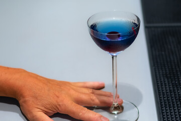 Male hand on bar counter next to stylish glass filled with deep blue famous cocktail Aviation with a single cherry