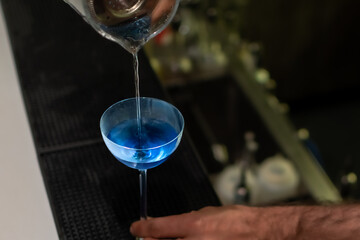 A bartender's hands are seen pouring a vibrant blue cocktail from cocktail shaker into stemmed coupe glass