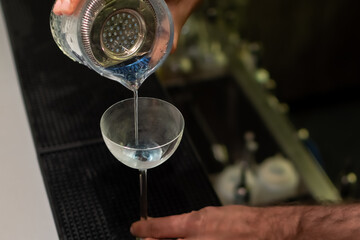 A close-up action shot of a bartender straining drink from mixing glass with julep strainer into...