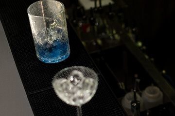 High-angle shot shows tall glass filled with vibrant blue liquid and ice, next to frosted coupe glass filled with ice on black bar mat