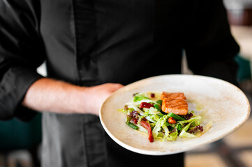 Chef in black uniform presenting grilled salmon with fresh salad and sauce on white plate, showcasing gourmet plating and culinary artistry