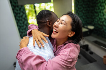 Obraz premium Excited child-free multiethnic couple embracing and laughing in modern living room, calm lifestyle