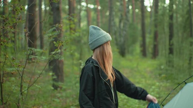 Young woman in black jacket and gray beanie holds tent cover in hand, smiling toward companion as she prepares to drape it over tent in peaceful forest with soft sunlight filtering through trees