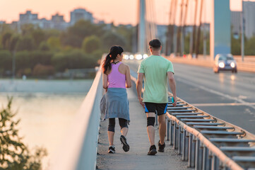 Asian woman and Caucasian man walking together on bridge at sunset after workout, enjoying conversation, showing friendship, diversity, and active healthy lifestyle