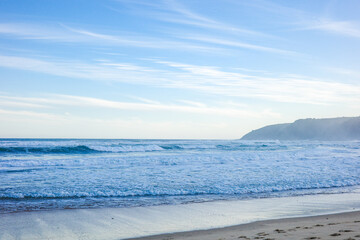 Obraz premium A calm seascape featuring gentle ocean waves rolling onto the sandy shoreline under a clear blue sky with wispy clouds and a misty coastal hill in the distance. The natural beauty of coastline