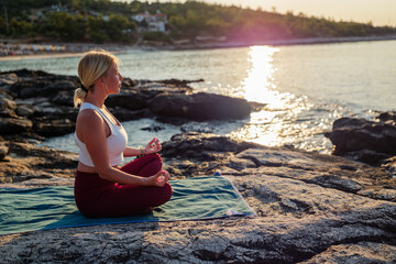 Young woman practicing yoga and meditating in lotus position on rocky beach at sunset, enjoying peace, mindfulness, and connection with nature