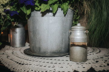 Obraz premium A cozy rustic display featuring metal buckets filled with fresh white and purple flowers, flanked by lanterns with lit candles, set on a lace-covered wooden cabinet.
