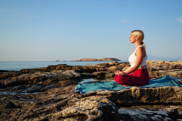 Young woman practicing yoga and meditation at sunrise on a rocky beach, embracing peace and...