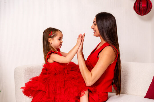 Happy mom in red suit and little daughter in fluffy dress sitting on sofa near Christmas tree, have fun and enjoying holidays. Parenthood and happy family concept. Merry Christmas and Happy New year