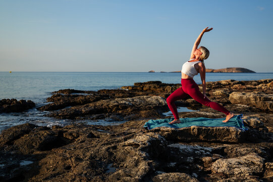 Woman performs a yoga warrior pose on a rocky beach at sunrise, finding peace and connection with nature as she stretches her body and embraces the tranquility of the ocean view