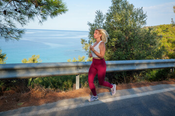 Young woman enjoys her outdoor workout, jogging along a scenic coastal road, embracing the breathtaking view of the turquoise sea and lush greenery