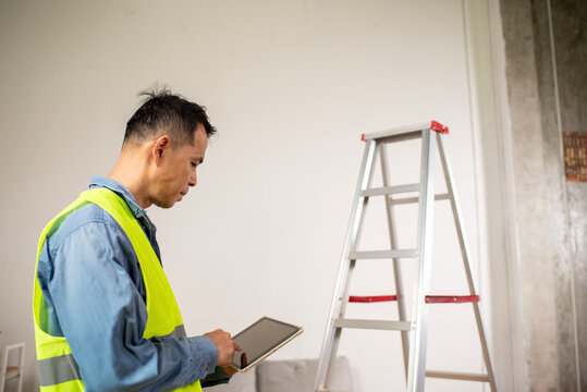 Construction Worker with Tablet: A focused construction worker wearing a high-visibility vest meticulously reviews a tablet near a ladder, poised amidst an unfinished indoor setting.