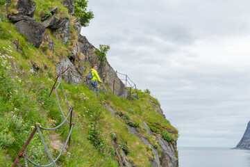 Caucasian hiker on Knuten mountain using nordic walking sticks. Rocks in foreground, steep mountain slope with grass and wild flowers. Ocean in the background. Cloudy summer day, Senja, Norway.