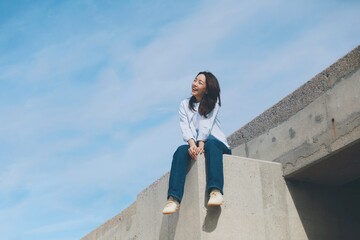 Cheerful young Asian woman sitting  on the roof under blue sky background.