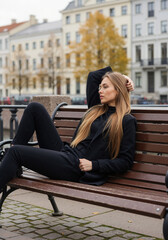 Fototapeta premium Woman in black coat lying on wooden bench with hand behind head in historic city square. Relaxed autumn elegance for luxury fashion brands, city lifestyle campaigns and sophisticated outerwear