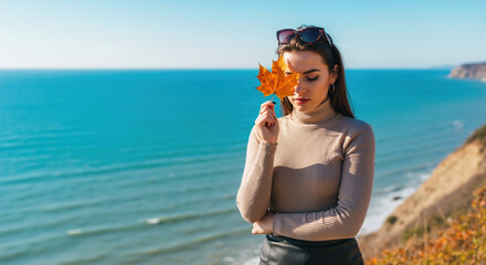 Woman in beige ribbed turtleneck with sunglasses holding orange maple leaf by coastal cliff. Coastal autumn style for travel fashion brands, knitwear campaigns and seaside lifestyle promotions