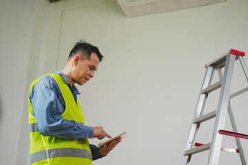 Construction worker wearing safety vest using tablet for project management while standing near ladder in modern workspace with empty wall © pichet
