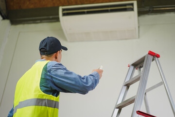 Technician Adjusts Air Conditioning Unit with Remote Control While Standing on Ladder in Industrial...
