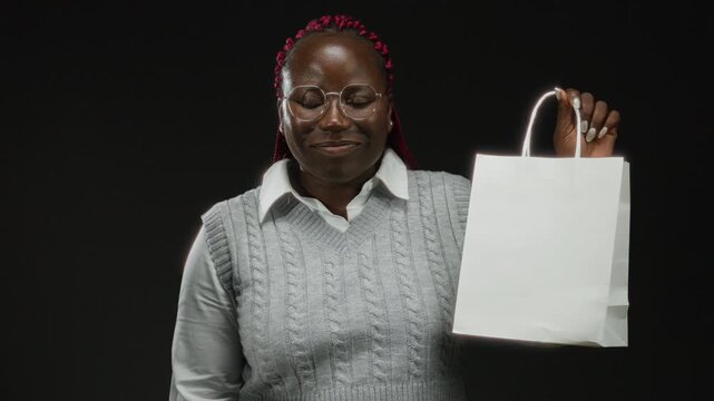 Woman in glasses with red braids holds a white shopping bag confidently against a black background, showcasing fashion and style with a modern and isolated demeanor.