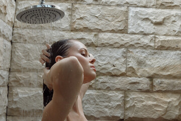 Woman taking shower under rainfall showerhead with natural stone wall background, relaxed...