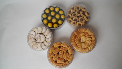 Five transparent plastic jars filled with: Nastar, Kastengel, Lidah Kucing, Putri Salju, and Chocolate Thumbprint Cookies. Each jar is neatly arranged in a row and isolated on a clean white background