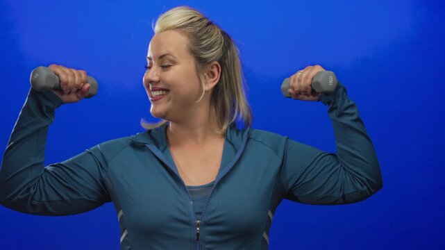 Young blonde woman in a sporty jacket smiling lifts 1kg dumbbells with both arms in a blue studio; strength motivation energy confidence.