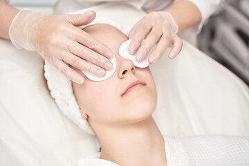 Young caucasian female receiving spa facial treatment with eye pads and gloved hands in relaxing setting.