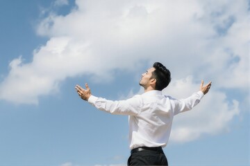 A man in a white shirt stands with arms outstretched against a bright blue sky with fluffy clouds.