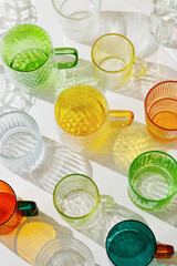 Vibrant collection of colorful glass cups on white background, casting artistic light reflections and shadows in natural sunlight. Summer drinks concept, minimal trend pattern of transparent glasses