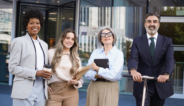 Diverse business team smiling together outside modern office building, one holding an electric scooter