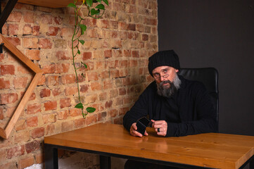 Confident modern businessman with a beard, sitting at a desk in a loft office with a brick wall,...