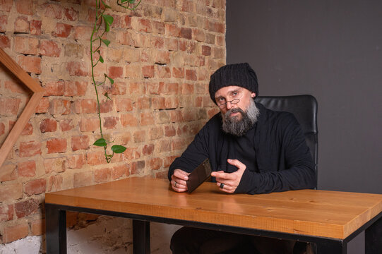 Portrait of a serious bearded hipster man in glasses and beanie sitting at a table with a smartphone. Creative freelancer in a loft office against a brick wall background