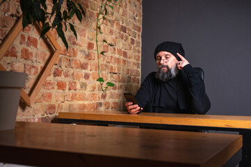 Confident modern businessman with a beard, sitting at a desk in a loft office with a brick wall,...