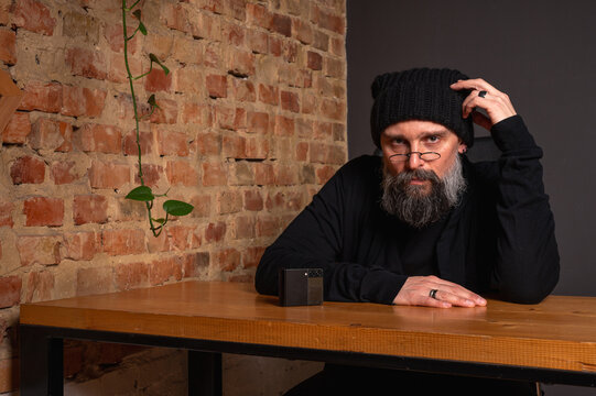 Portrait of a serious bearded hipster man in glasses and beanie sitting at a table with a smartphone. Creative freelancer in a loft office against a brick wall background - Powered by Adobe