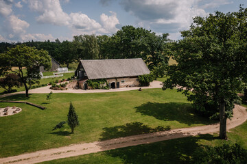 A large rustic countryside barn with a gray roof surrounded by a green lawn, gravel paths, trees, and a peaceful garden on a sunny day with scattered clouds.