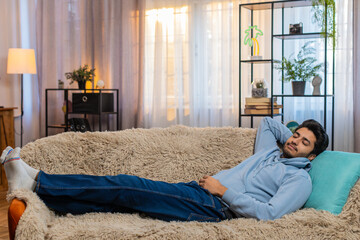 Tired Indian man lying down on comfortable couch, taking rest, daytime nap. Carefree Hindu guy relaxing, falling asleep at home. Hispanic adult male resting with eyes closed, looking calm and serene.