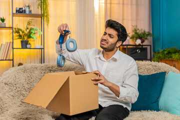 Sad Indian young man at home opening a package, shocked to find damaged wireless headphones inside....
