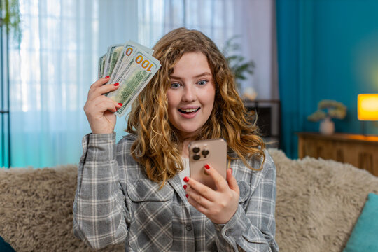Young woman at home smiling after receiving victory SMS on smartphone about incoming cash dollars feeling happy. Caucasian redhead girl on sofa showing thumbs up, joyful reaction to financial success