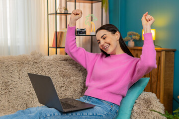 Young woman at home raising hands up with excitement and joy celebrating successfully completed...