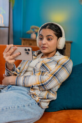 Indian young woman relaxing at home on sofa, wearing headphones and happily listening to podcast or...