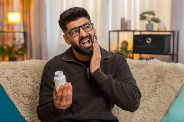 Indian man at home holds cheek with painful face looking for painkillers due to strong dental discomfort. Arabian guy on sofa reacts to toothache feeling unwell and stressed by aching health condition